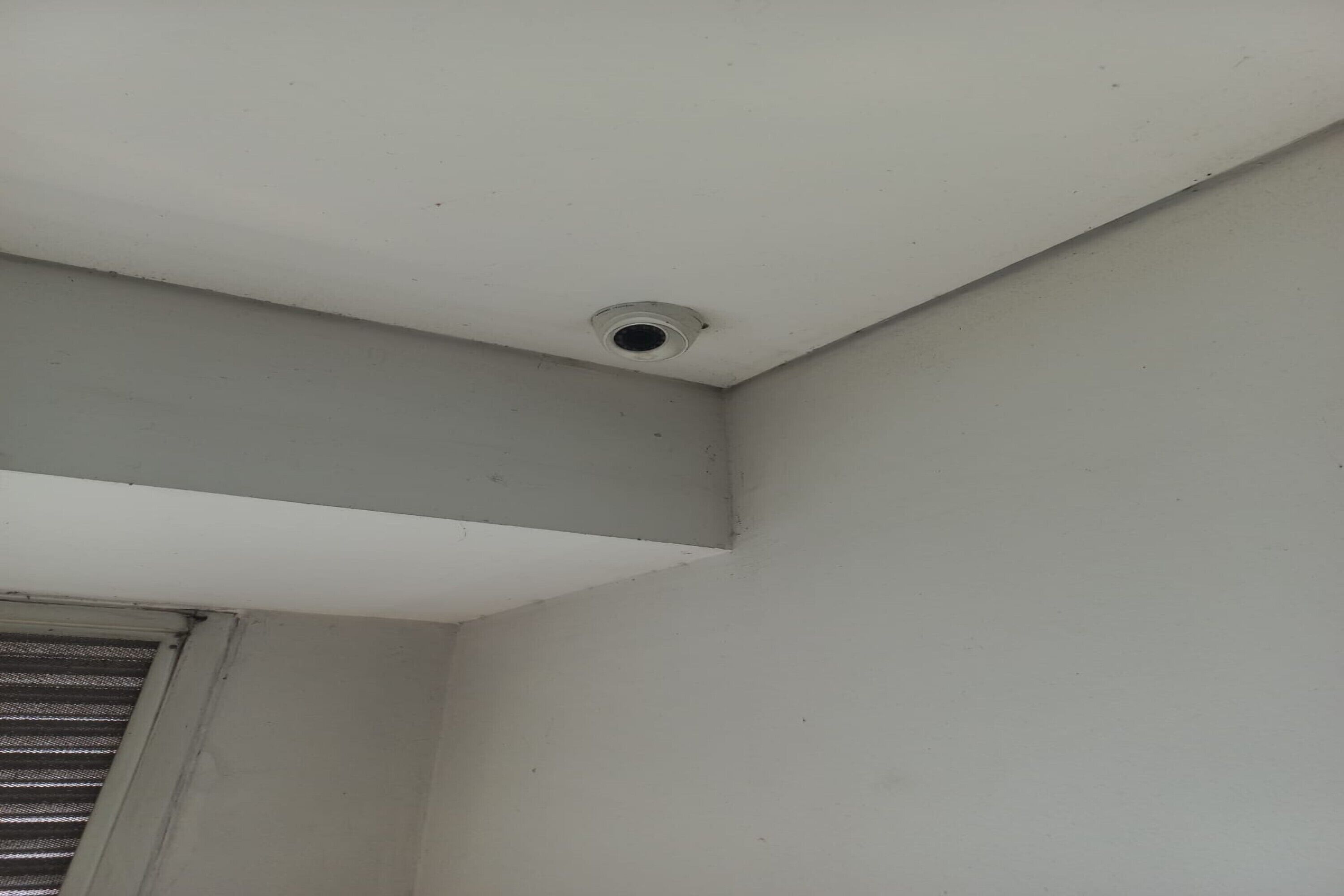 A corner of a room with a ceiling vent and a window with horizontal blinds.