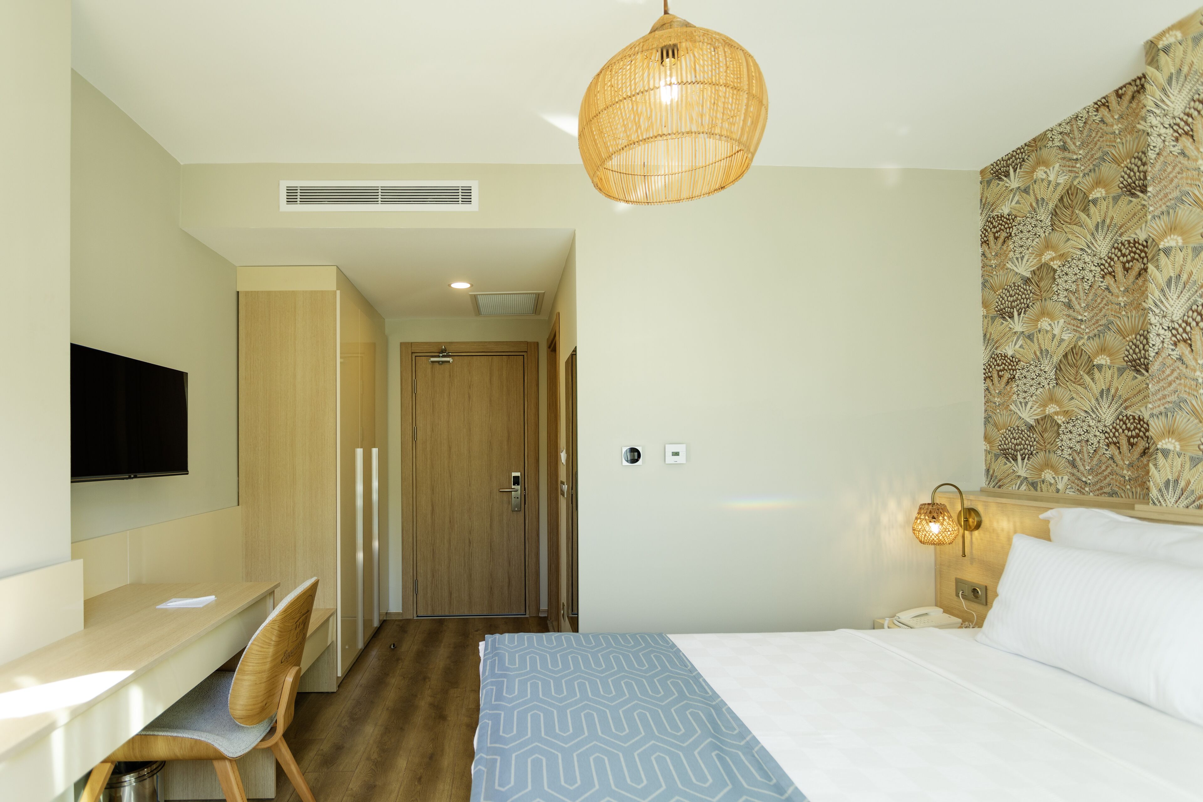 A hotel room with a bed, a desk, a chair, a television, and a wall-mounted light fixture.