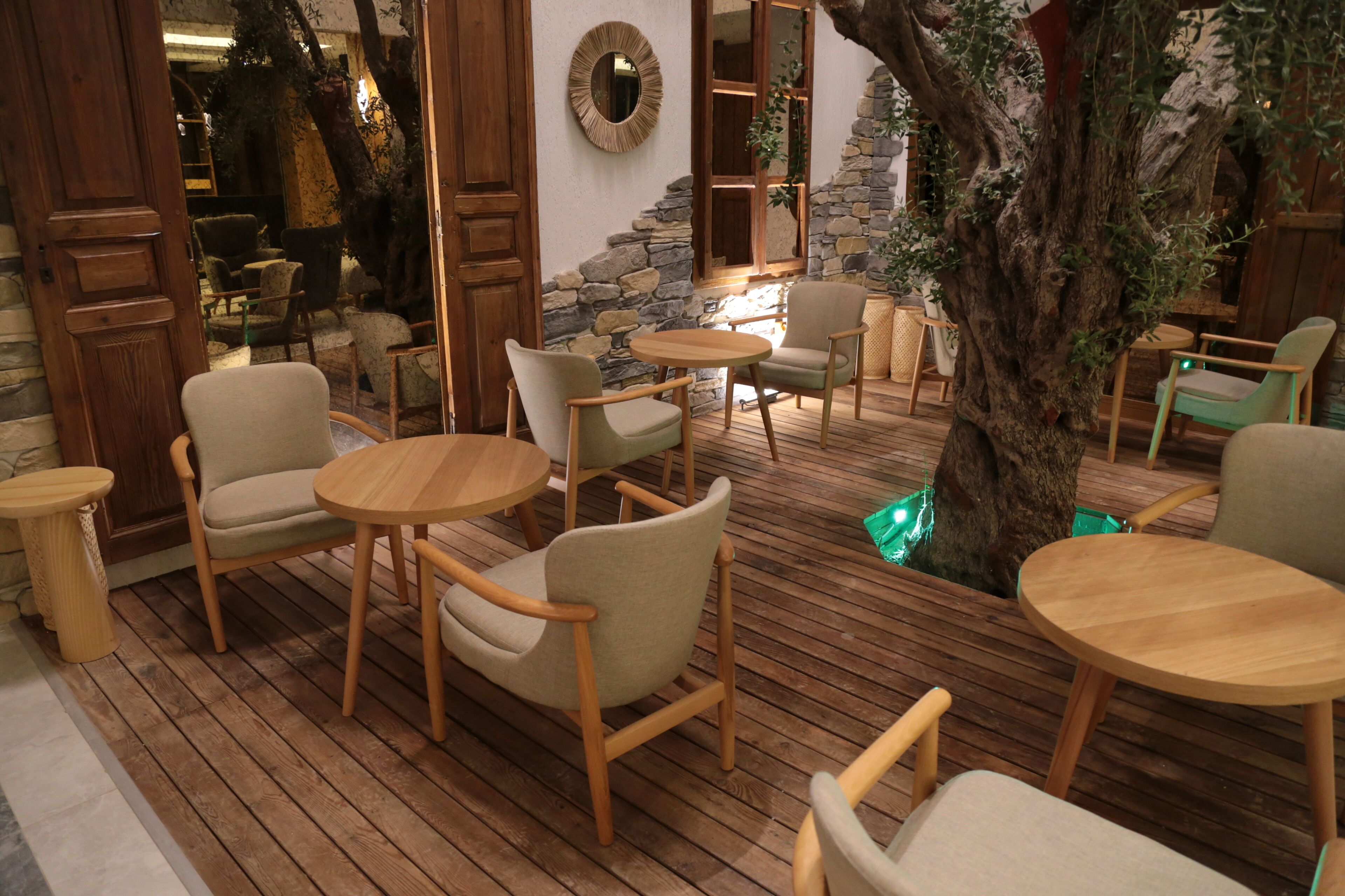 Un área de estar interior acogedora, con mesas y sillas de madera, rodeada de paredes de piedra y un gran árbol.
