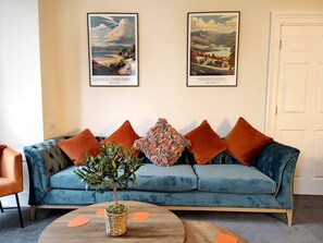 Un salon avec un canapé de couleur turquoise, des coussins orange, une petite plante en pot et deux affiches représentant des paysages, encadrées et accrochées au mur.