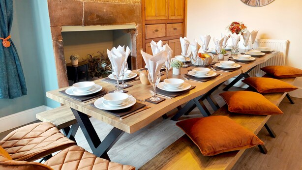Un coin repas avec une longue table en bois dressée pour le repas, entourée de chaises rembourrées avec des coussins orange.