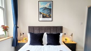 Un lit bien fait, recouvert d’une couverture bleue et agrémenté de deux oreillers, une photo encadrée du Scafell Pike, et une fenêtre avec des rideaux bleus.
