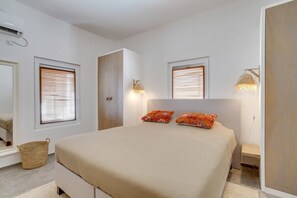 Un dormitorio con una cama, dos lámparas de noche, un ventanal con persianas, un aire acondicionado de pared y una pequeña cesta de mimbre.