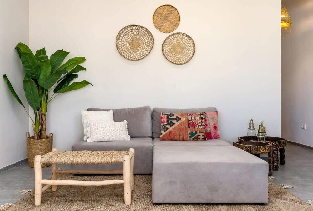 A modern living room with a grey sofa, a wooden bench, and a grey ottoman. There are two decorative wall hangings above the sofa and a potted plant to the left.