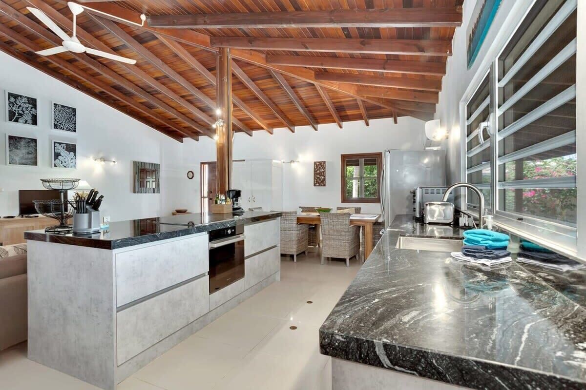 A modern kitchen with a central island, wooden ceiling beams, and a dining area with a view of greenery.