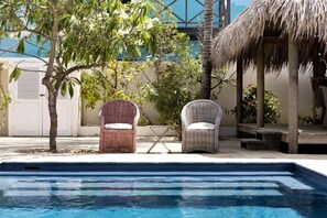 Un área de piscina con sillas de mimbre, una estructura de paja y un árbol.