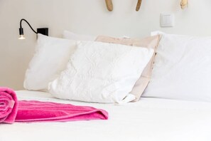 En pænt redt seng med hvide sengetøj og et pink håndklæde, samt en vægmonteret lampe og en stikkontakt.