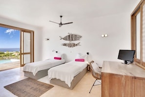 A modern bedroom with two beds, a desk, a chair, a ceiling fan, and a view of the ocean. - Kas Berde with lap pool and sea views (Kralendijk)