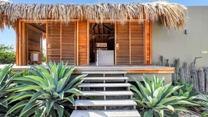 A wooden building with a thatched roof, a wooden staircase leading to a door, and large green plants in the foreground. - Kas Berde with a large private pool and spectacular view towards Klein Bonaire (Kralendijk)