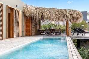 A pool area with a wooden deck, a thatched roof structure, and lounge chairs.