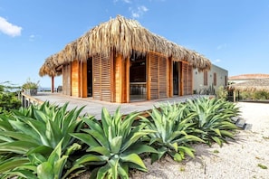 A thatched-roof building with wooden shutters, surrounded by a wooden deck and large green plants. - Kas Berde with lap pool and sea views (Kralendijk)