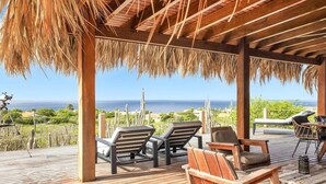 A wooden deck with lounge chairs and a table, overlooking a coastal landscape with cacti and a clear view of the ocean. - Kas Berde with a large private pool and spectacular view towards Klein Bonaire (Kralendijk)