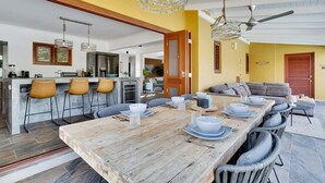 A spacious dining area with a wooden table set for six, surrounded by wicker chairs. A kitchen with a bar area is visible in the background. - Kas Pura Vida with a very cozy poolside palapa (Kralendijk)