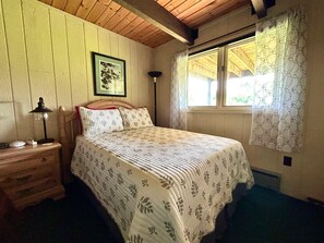 Quarto com cabeceira de madeira, mesa de cabeceira com abajur, quadro emoldurado na parede e janela com cortinas estampadas.