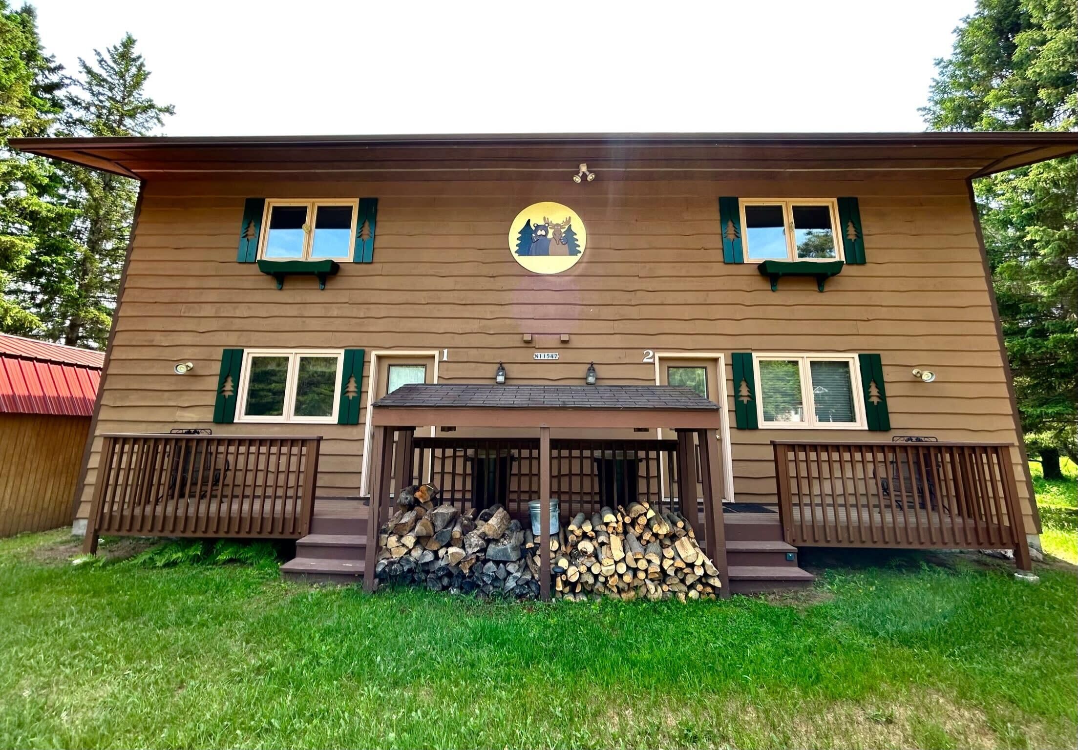 A two-story cabin with a central circular emblem, two windows with shutters, and a covered porch with stacked firewood.
