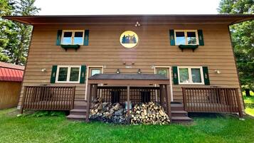 A two-story cabin with a central circular emblem, two windows with shutters, and a covered porch with stacked firewood.