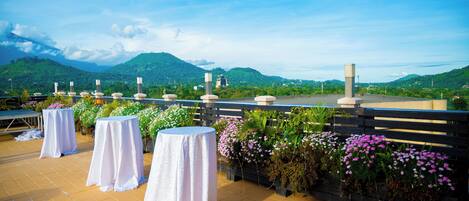 A terrace with tables covered in white tablecloths, surrounded by potted plants and flowers, overlooking a mountainous landscape.