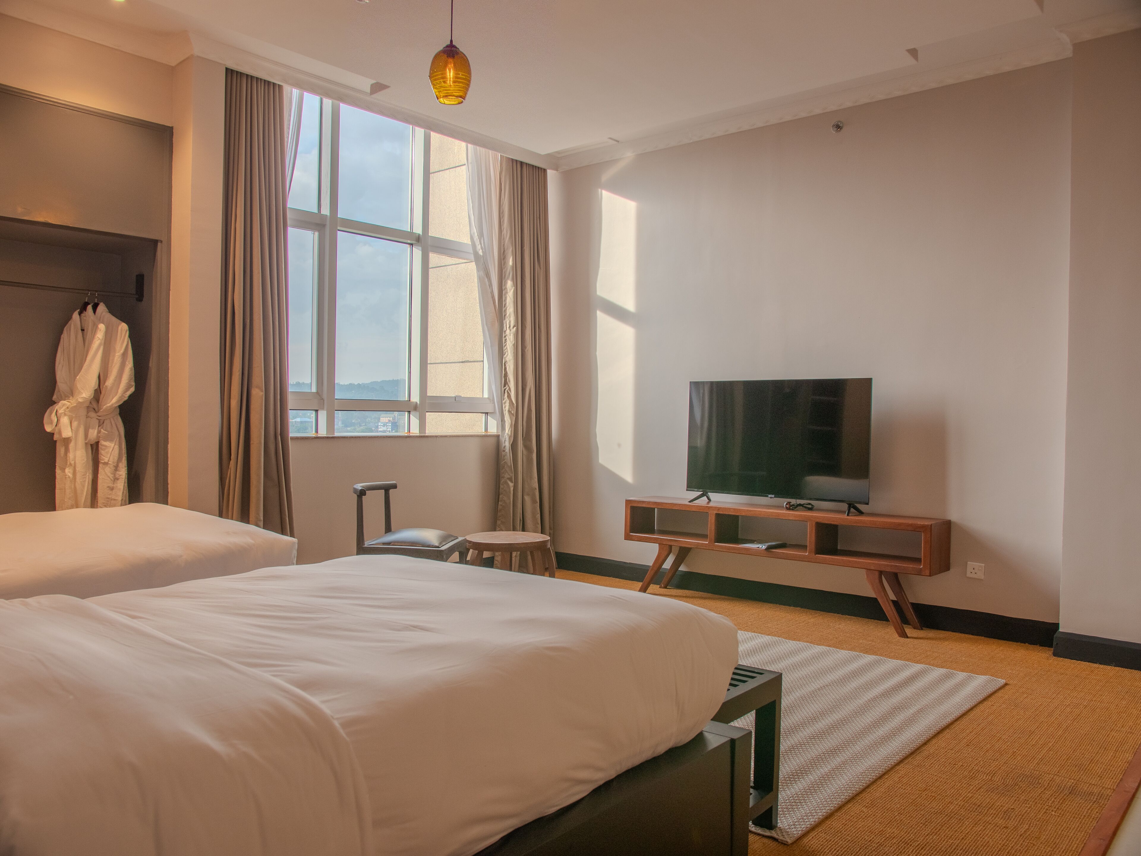 Habitación de hotel con dos camas, un televisor sobre un soporte de madera, una bata colgada en un perchero y vistas al exterior a través de amplios ventanales.