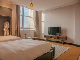 Habitación de hotel con dos camas, un televisor sobre un soporte de madera, una bata colgada en un perchero y vistas al exterior a través de amplios ventanales.