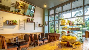 A restaurant with wooden tables and chairs, a glass display case with pastries, and a large window with a view of trees. - Sanna Boutique Hotel (Arusha)