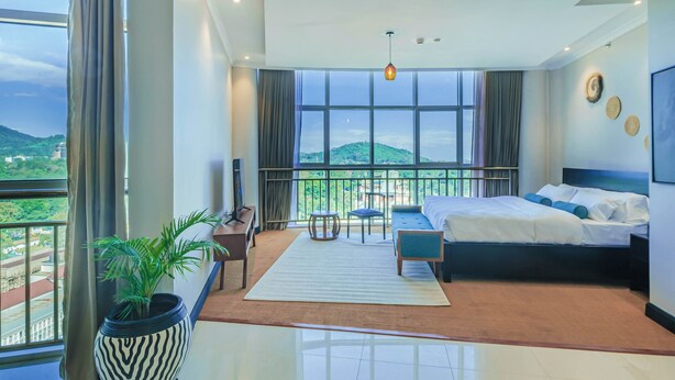 A modern bedroom with a large bed, a desk, a chair, and a view of a green landscape through large windows. - Sanna Boutique Hotel (Arusha)