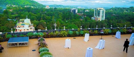 A rooftop terrace with a stage, white tables, and a view of a green hillside with buildings.