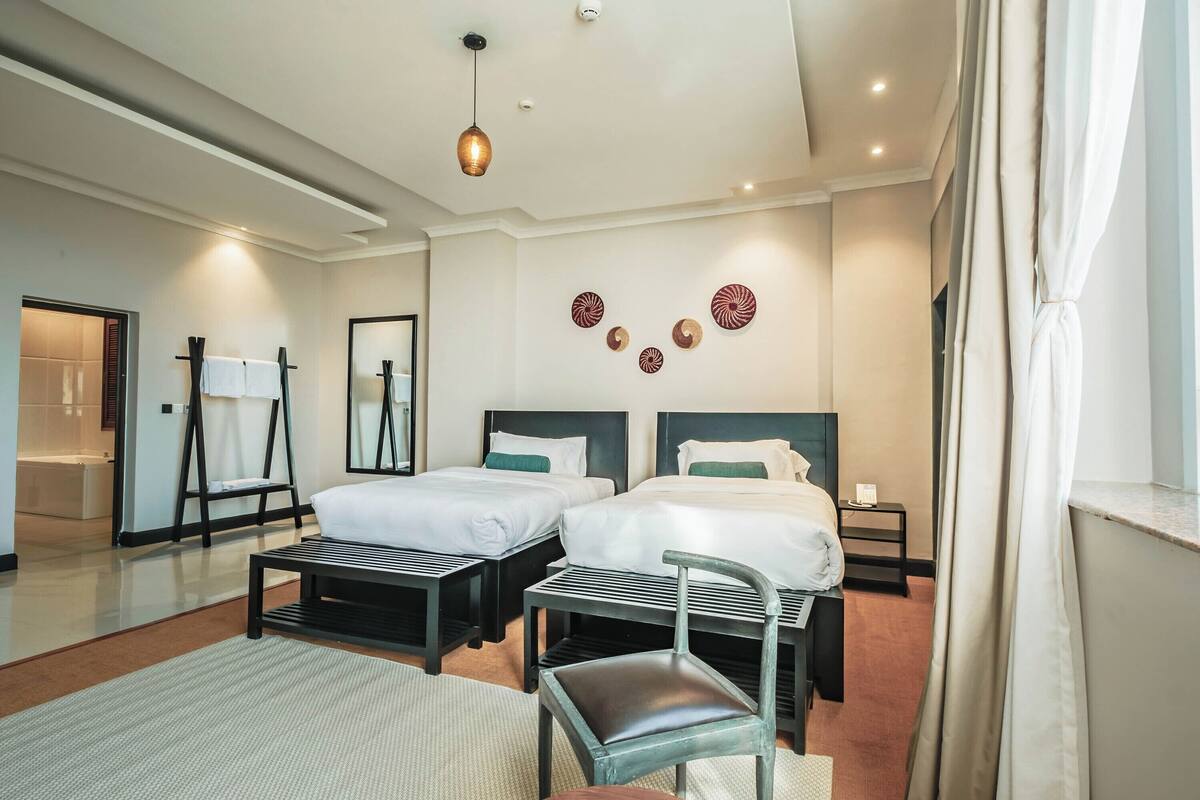 A modern hotel room with two beds, a small table, a chair, and a mirror.