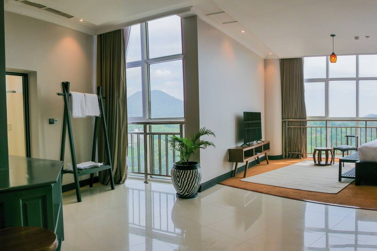 A modern hotel room with a large bed, a desk with a chair, a TV, and a view of the mountains through large windows.