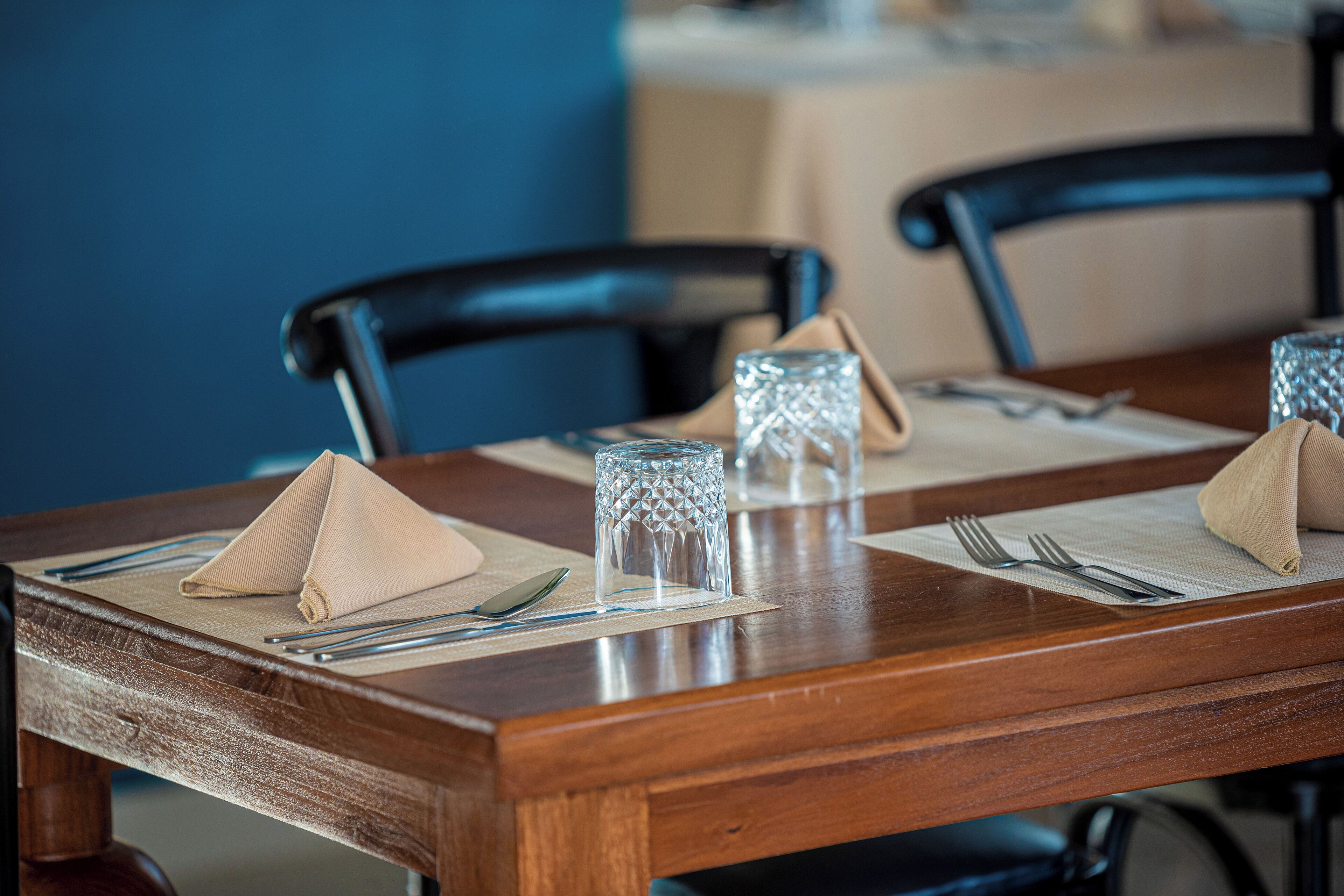 Een houten eettafel gedekt met beige servetten, bestek en helder glaswerk.