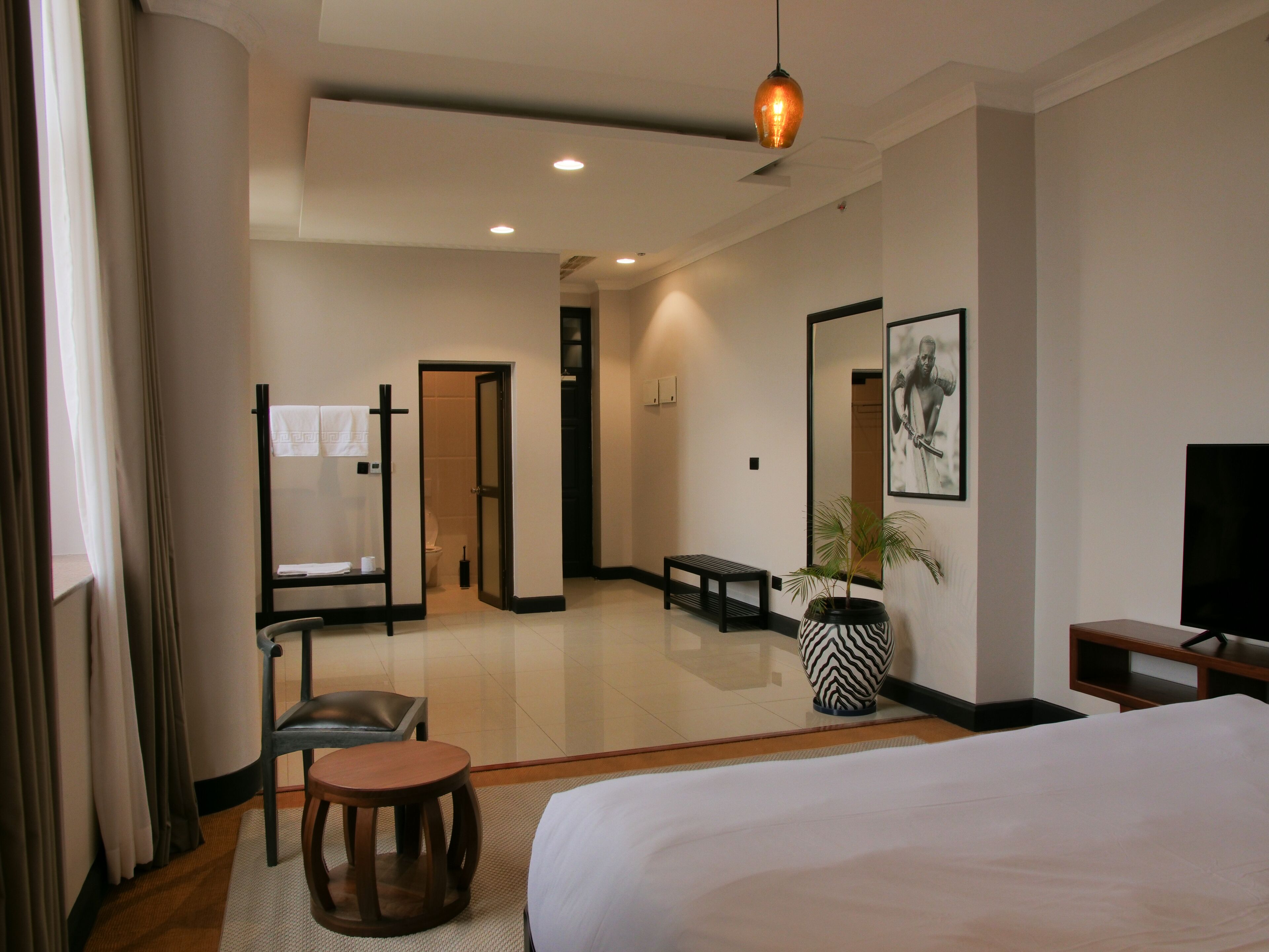 A hotel room with a bed, a desk, a chair, a small table, a plant, a bench, a TV, and a bathroom visible through an open door.