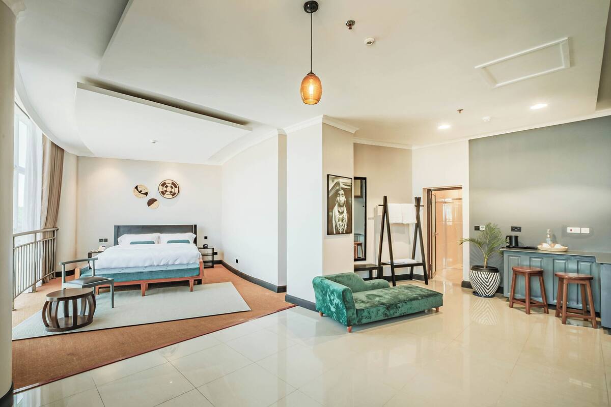 A modern hotel room with a large bed, a green sofa, a small table, and a kitchen area with stools.