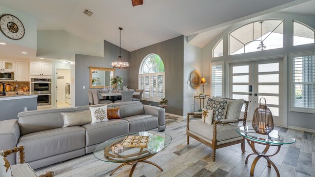 A spacious living room with a grey sofa, a glass coffee table, and a wooden chair with a glass top. A large window provides natural light and a view of the outdoors. - Gorgeous Punta Gorda Home w/ Lanai & Private Pool! (Punta Gorda)