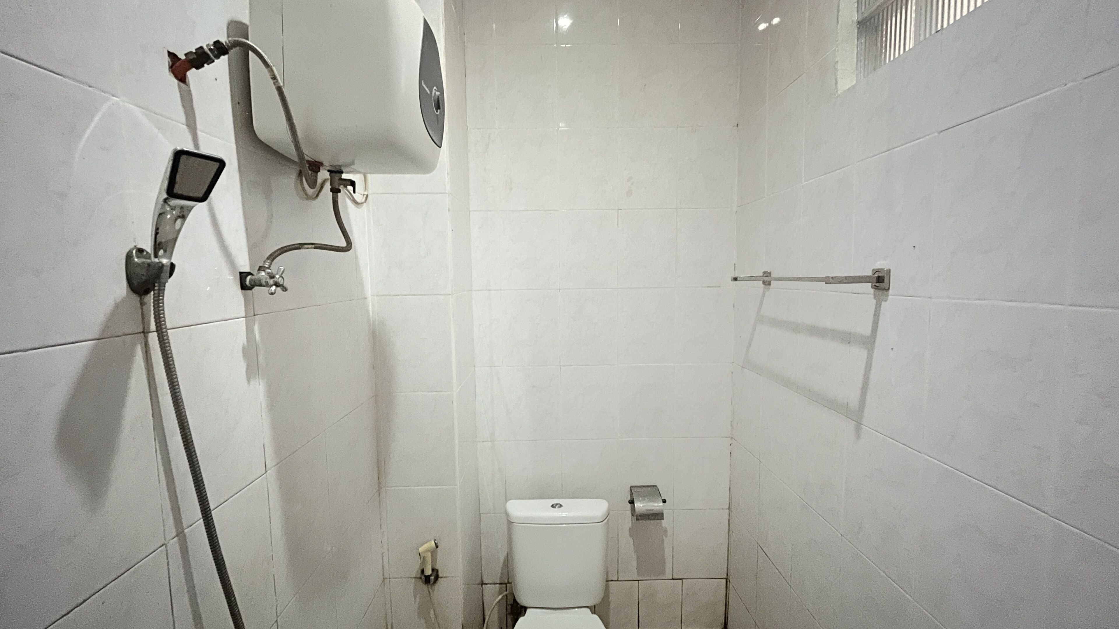 Un baño pequeño con azulejos, un inodoro, un cabezal de ducha de mano y un perchero para toallas.