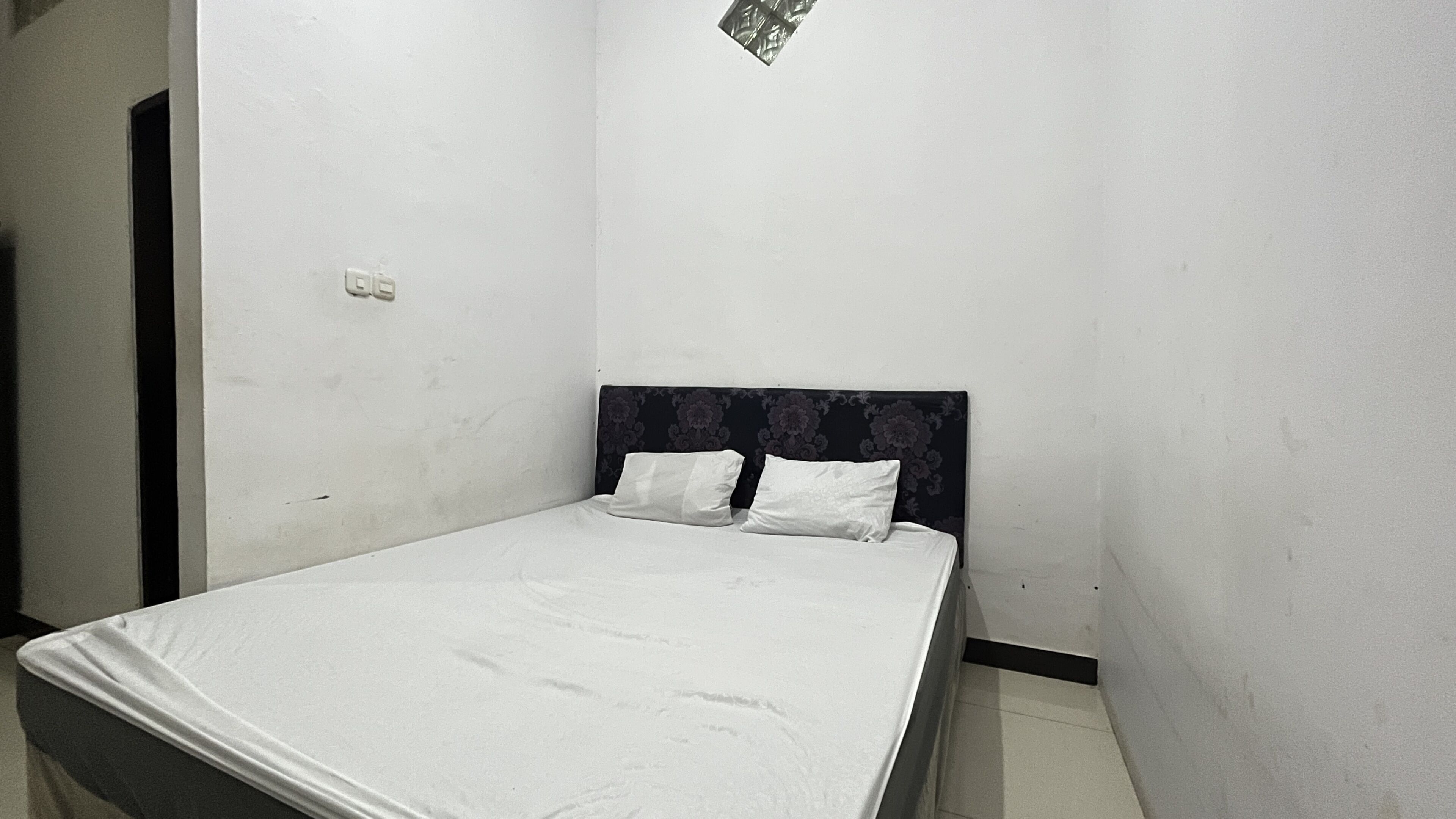 Una habitación pequeña con una cama individual, una colcha blanca sencilla y dos almohadas. El cabecero tiene un estampado floral, y hay una puerta pequeña de color oscuro en el lado izquierdo.