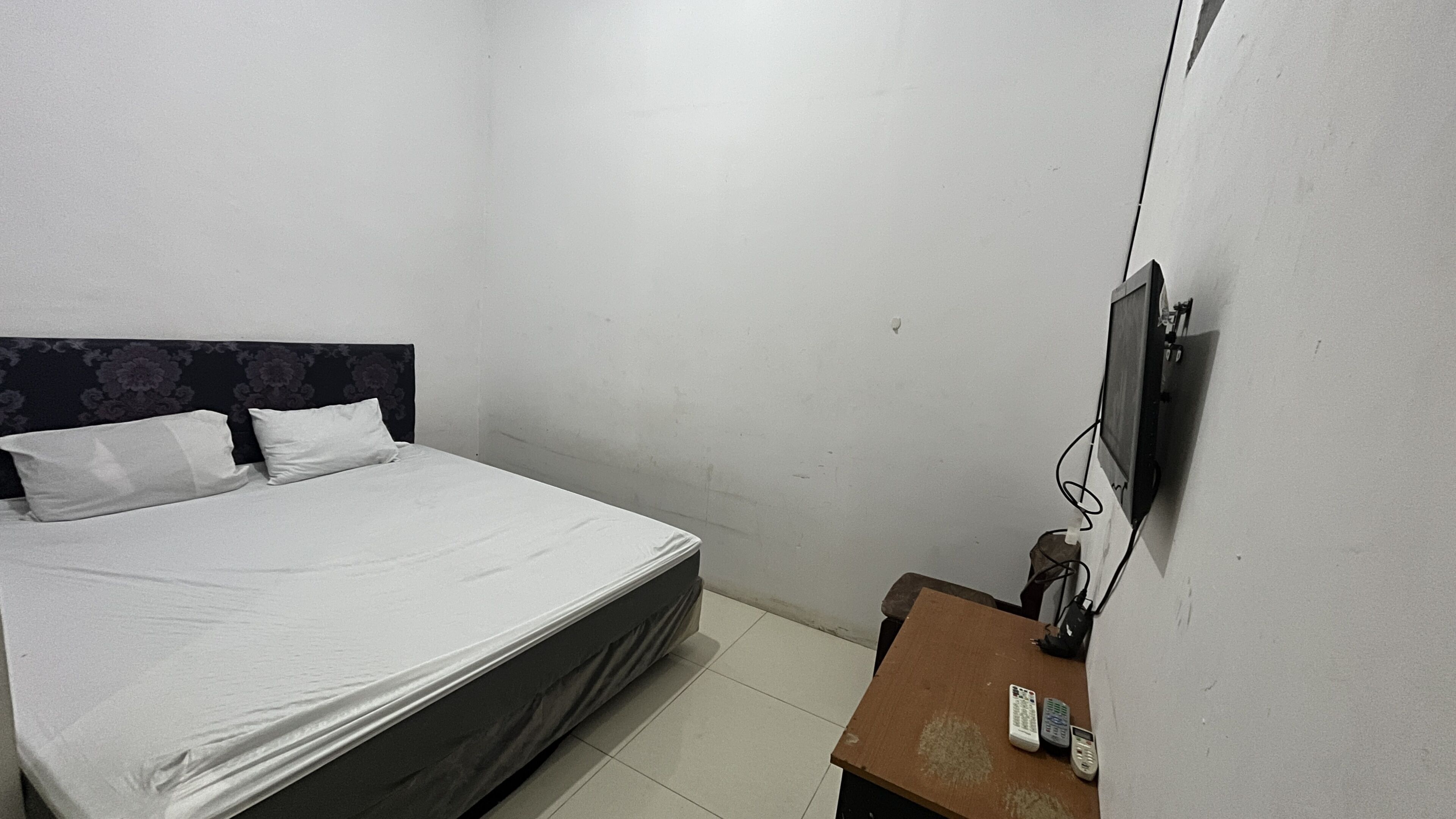 Una cama individual con cabecera, un televisor de pantalla plana montado en la pared y una mesita de noche de madera con dos controles remotos.