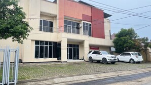 A multi-story building with a red and beige facade, large windows, and a sign reading "TERIMA KOST". Two white SUVs are parked in front. - Hotel O Guest House Sae Madani (Sepaku)