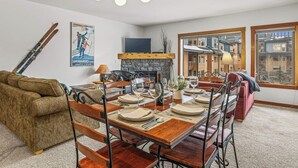 Un coin repas avec une table en bois dressée pour quatre personnes, une cheminée et une vue sur un bâtiment par la fenêtre.