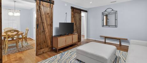 A room with a wooden floor, a television on a wooden cabinet, a ceiling fan, a bench, and a dining area with a table and chairs.