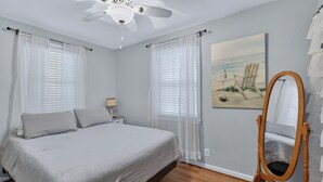 A bedroom with a bed, a ceiling fan, a painting of a beach scene, a wooden-framed mirror, and a nightstand with a lamp.