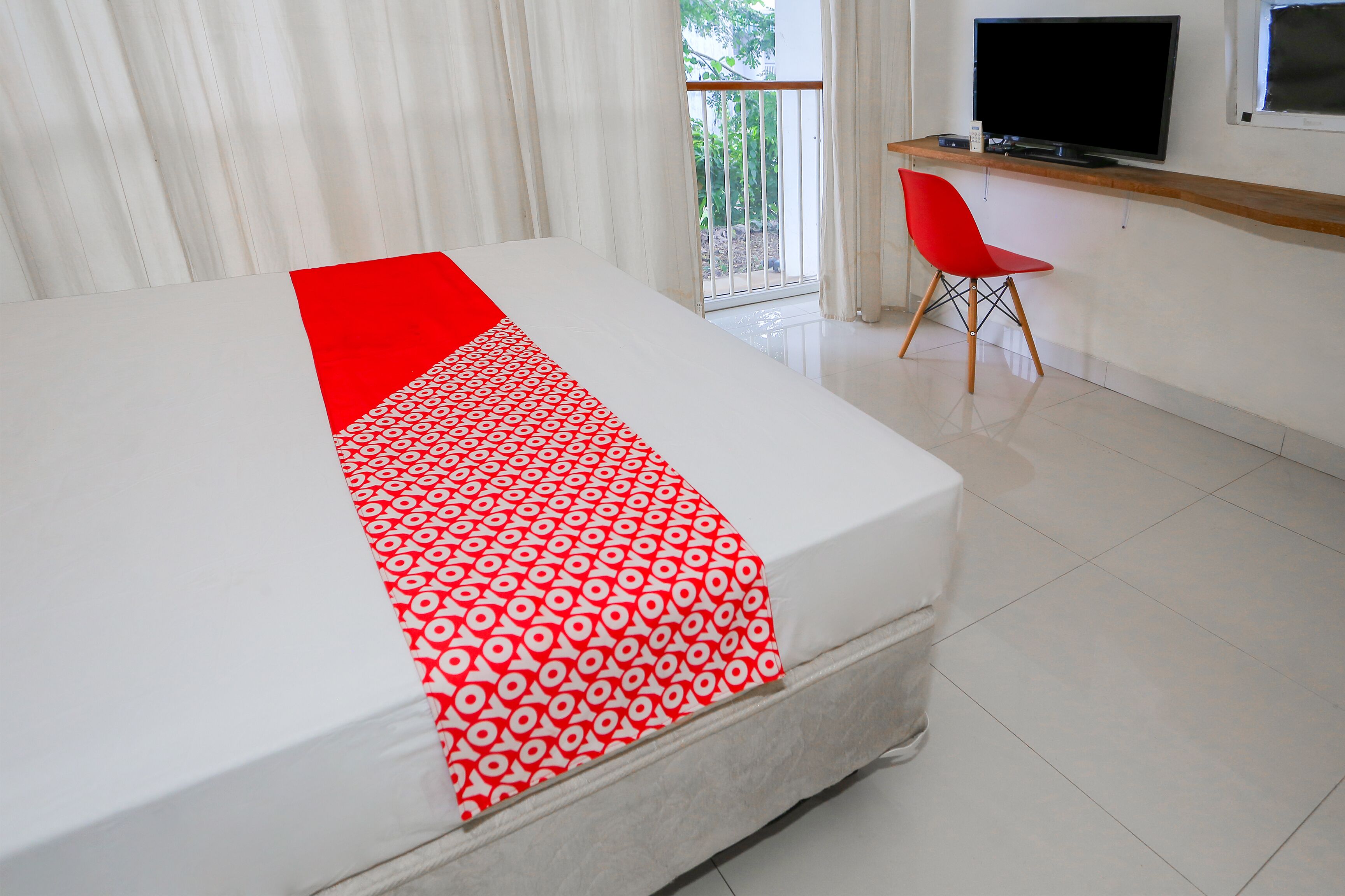 A hotel room with a bed, a television, a red chair, and a balcony with curtains.