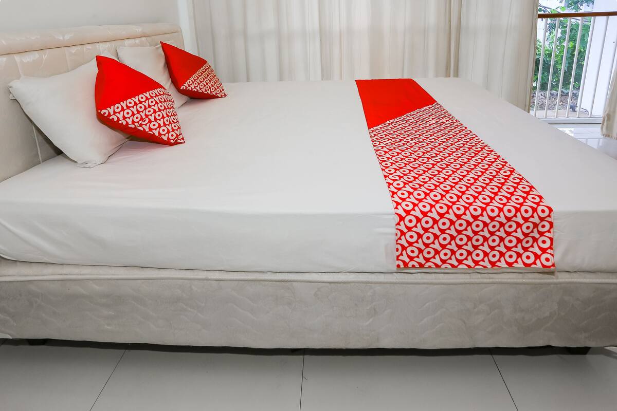 A bed with white and red bedding, a white headboard, and a view of greenery outside.
