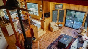 A spacious living area with a fireplace, a television, and a large window overlooking a deck. - Oasis | Ocean Views & Private Hot Tub (Sea Ranch)