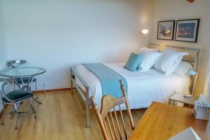 A bedroom with a bed, a bedside table, a chair, a small table, and a wall-mounted lamp. - Oasis | Ocean Views & Private Hot Tub (Sea Ranch)