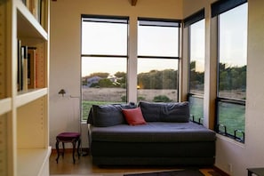 A room with a large window, a sofa, a small table, and a bookshelf. - Oasis | Ocean Views & Private Hot Tub (Sea Ranch)