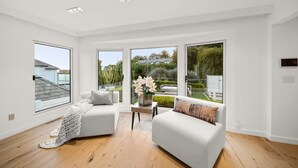 Un salon moderne avec deux canapés blancs, une petite table d’appoint sur laquelle est posé un vase de fleurs, et de grandes fenêtres donnant sur un jardin et les maisons voisines.