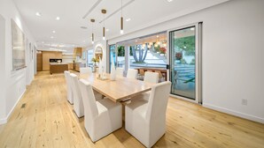 Un espace repas moderne avec une table en bois et des chaises blanches, une cuisine en arrière-plan et une grande porte-fenêtre coulissante donnant sur un espace extérieur.
