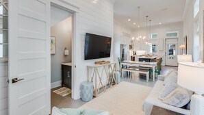 A spacious living room with a large flat-screen TV, a dining area, and a kitchen with a 'BEACH HOUSE' sign.