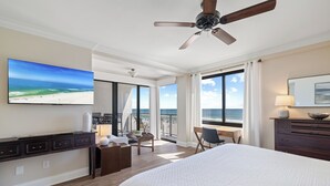 Quarto de hotel com uma cama grande, uma escrivaninha, uma cadeira, uma televisão, um ventilador de teto e uma varanda com vista para o oceano.