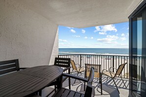 Ein Balkon mit einem Holztisch und Stühlen, der auf einen Strand mit Personen und Sonnenschirmen blickt.