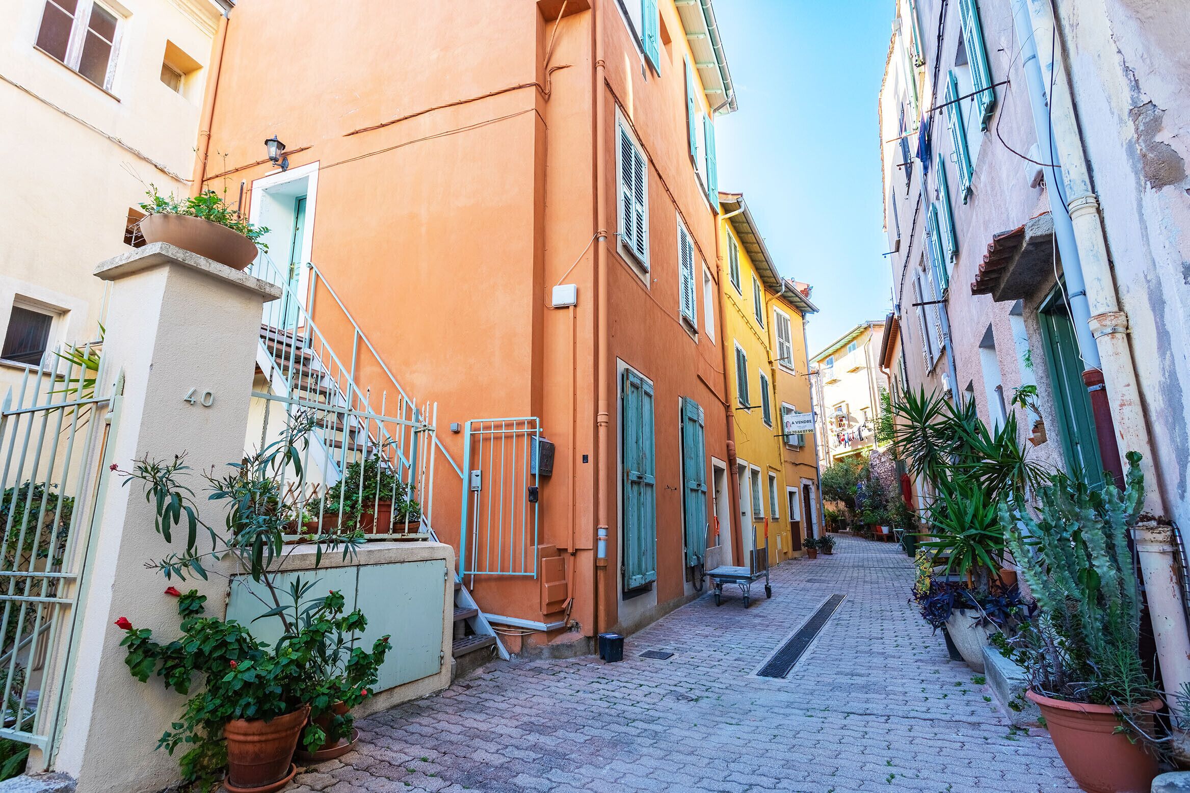 Une ruelle étroite pavée, bordée de bâtiments aux couleurs vives, de plantes en pot et d’un portail en métal.